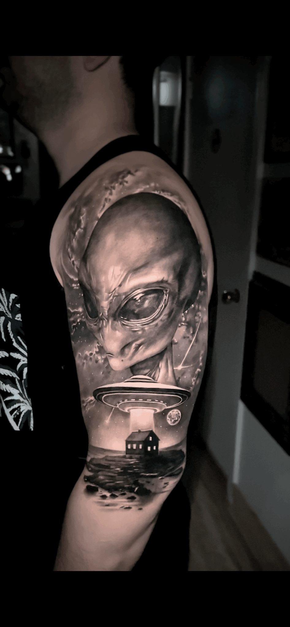 Tattoo of an alien face with large eyes on person's upper arm, with a boat and water scene below.