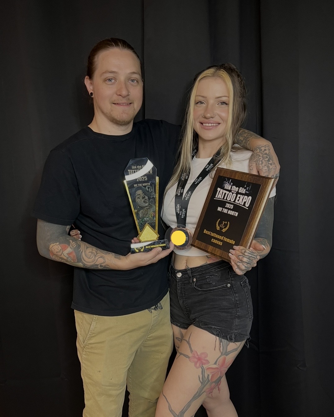 Two people standing together, one holding a trophy and a framed certificate, smiling, against a dark background.