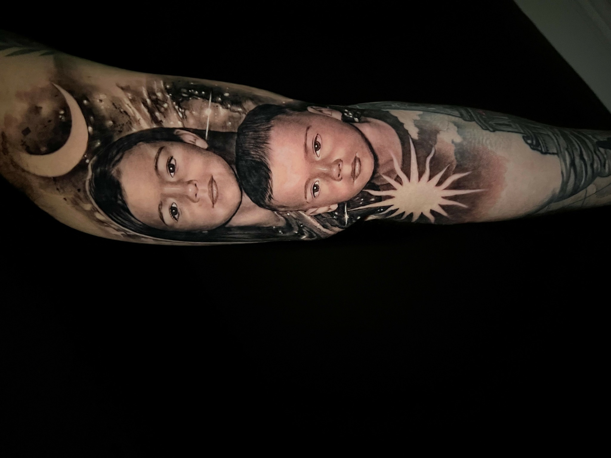 Tattoo of three faces, celestial bodies, and a sunburst design on a person's arm.