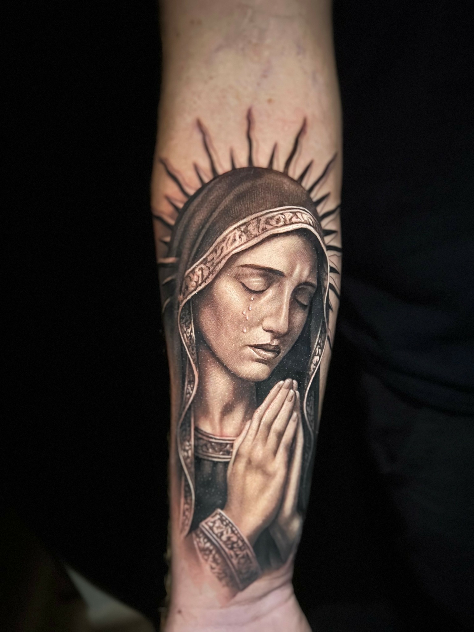 Tattoo of a woman with closed eyes and hands in prayer, surrounded by a halo with rays, on arm.