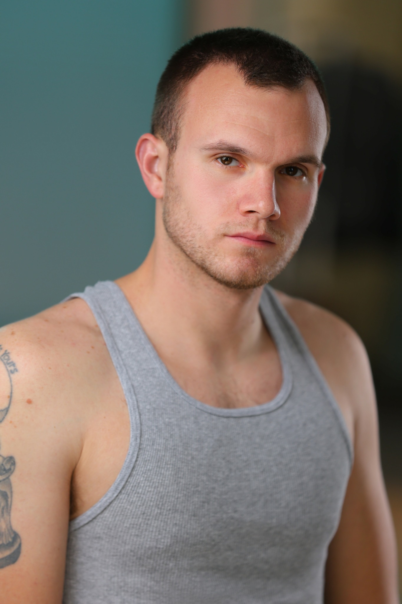 Young man with short dark hair and a serious expression, wearing a gray tank top, standing indoors.