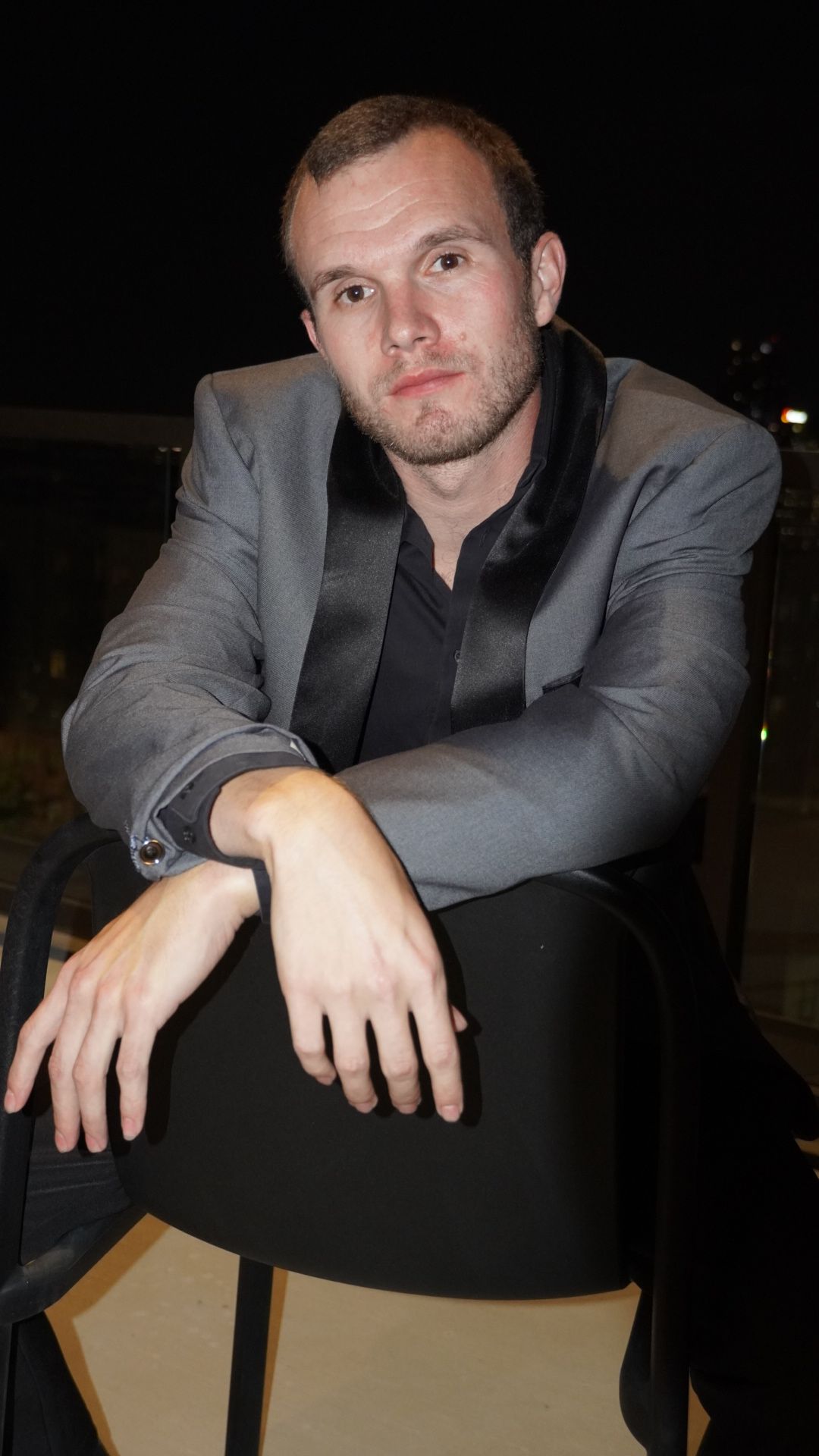 Man in gray suit and black tie sitting at a table, looking at the camera, with a neutral background.