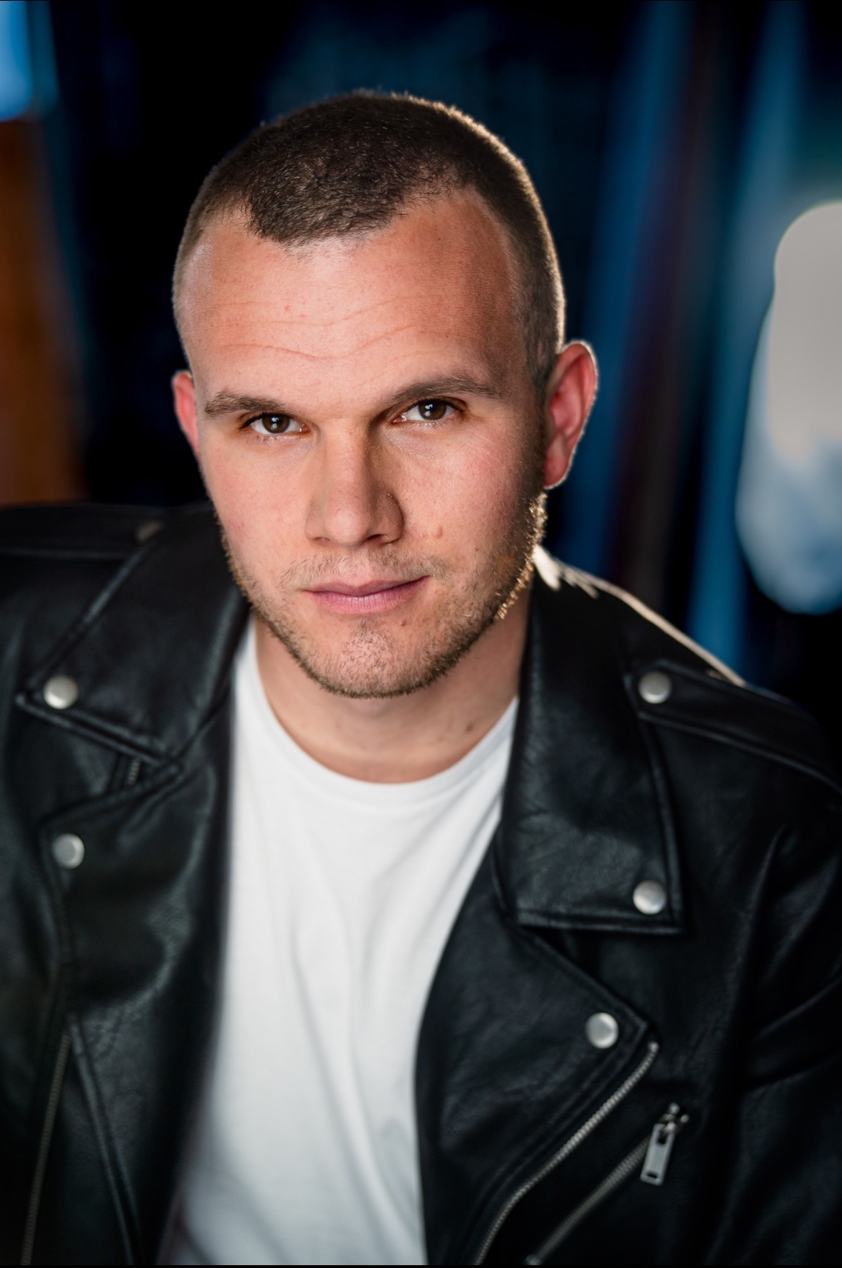Young man with short hair wearing a black leather jacket and white shirt, looking at camera.