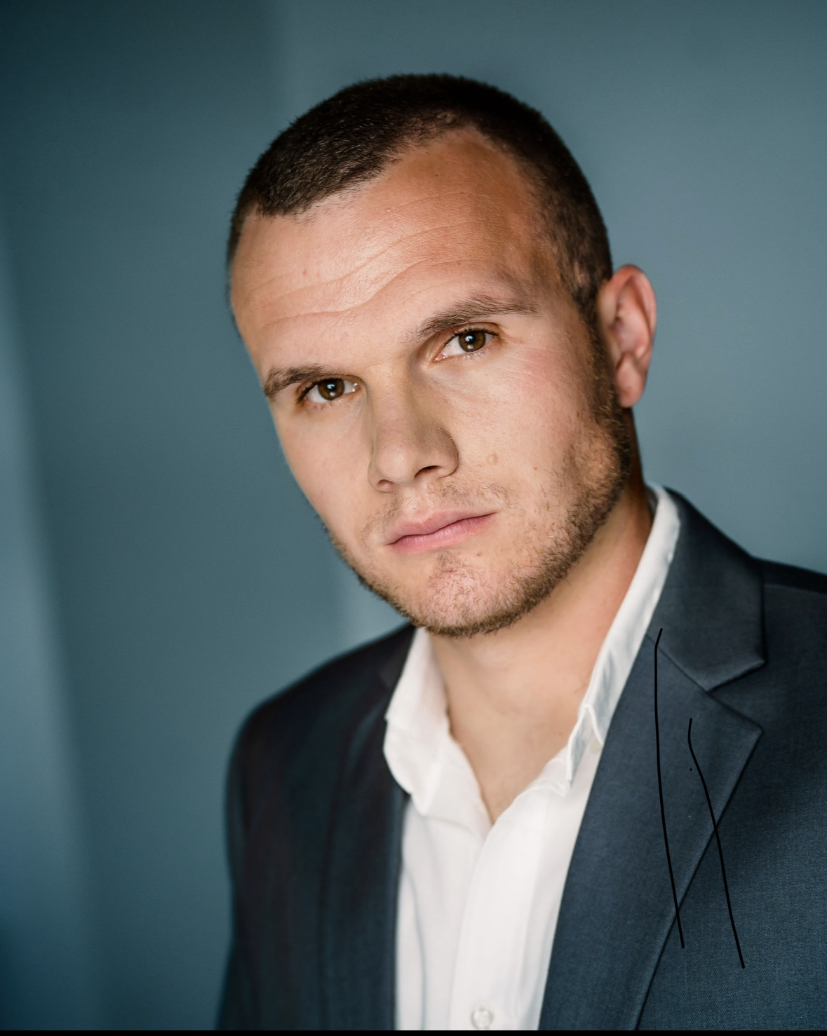 Portrait of a man with short hair, wearing a dark suit and white shirt, against a plain background.