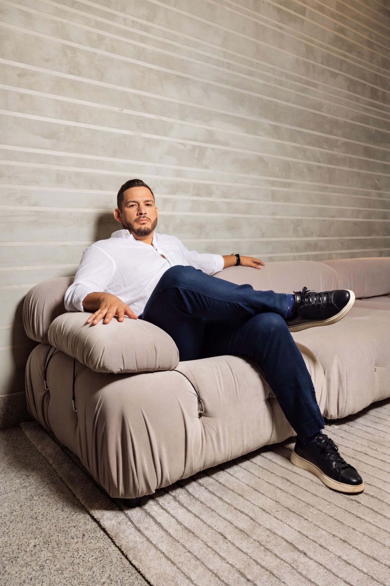 Man sitting on a beige sofa with a light-colored brick wall behind him, relaxed pose, wearing a white shirt and dark pants.