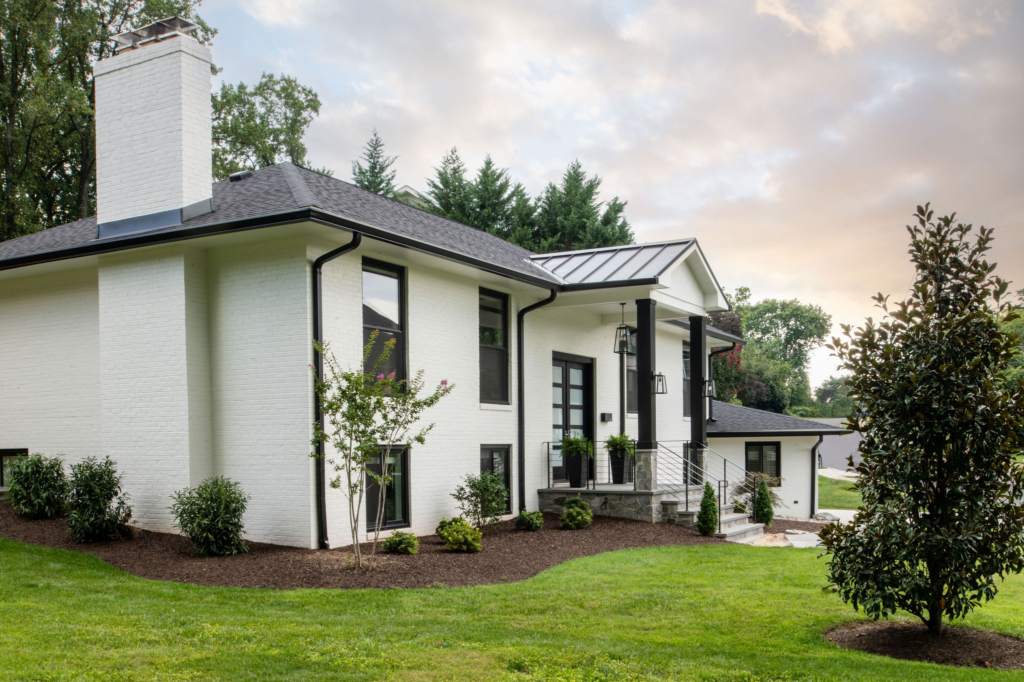 White house with black roof, surrounded by green lawn and trees, under a partly cloudy sky.