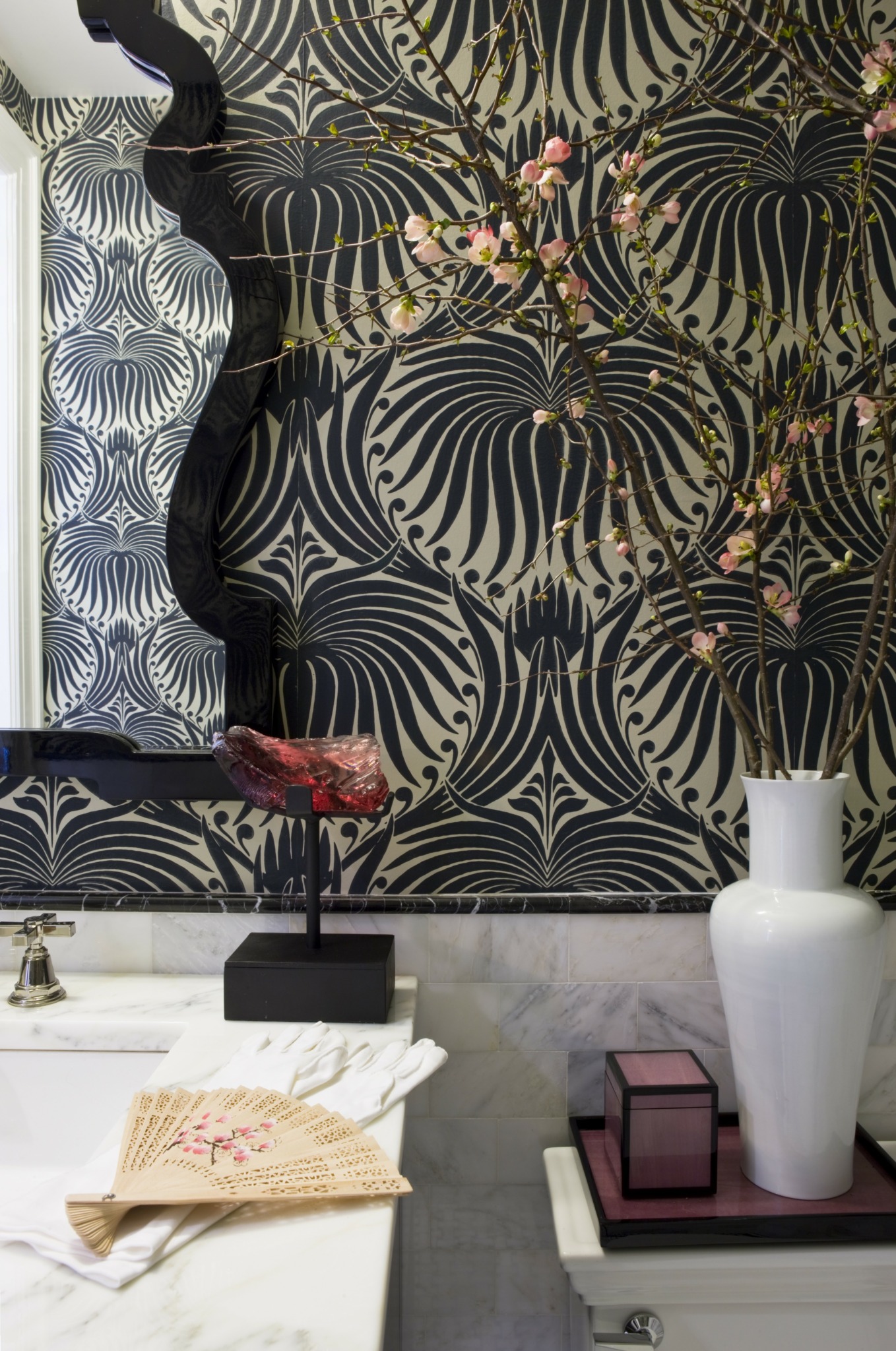 Decorative wall with black and gold floral pattern, white vase, pink box, and a table with a fan and cloths.