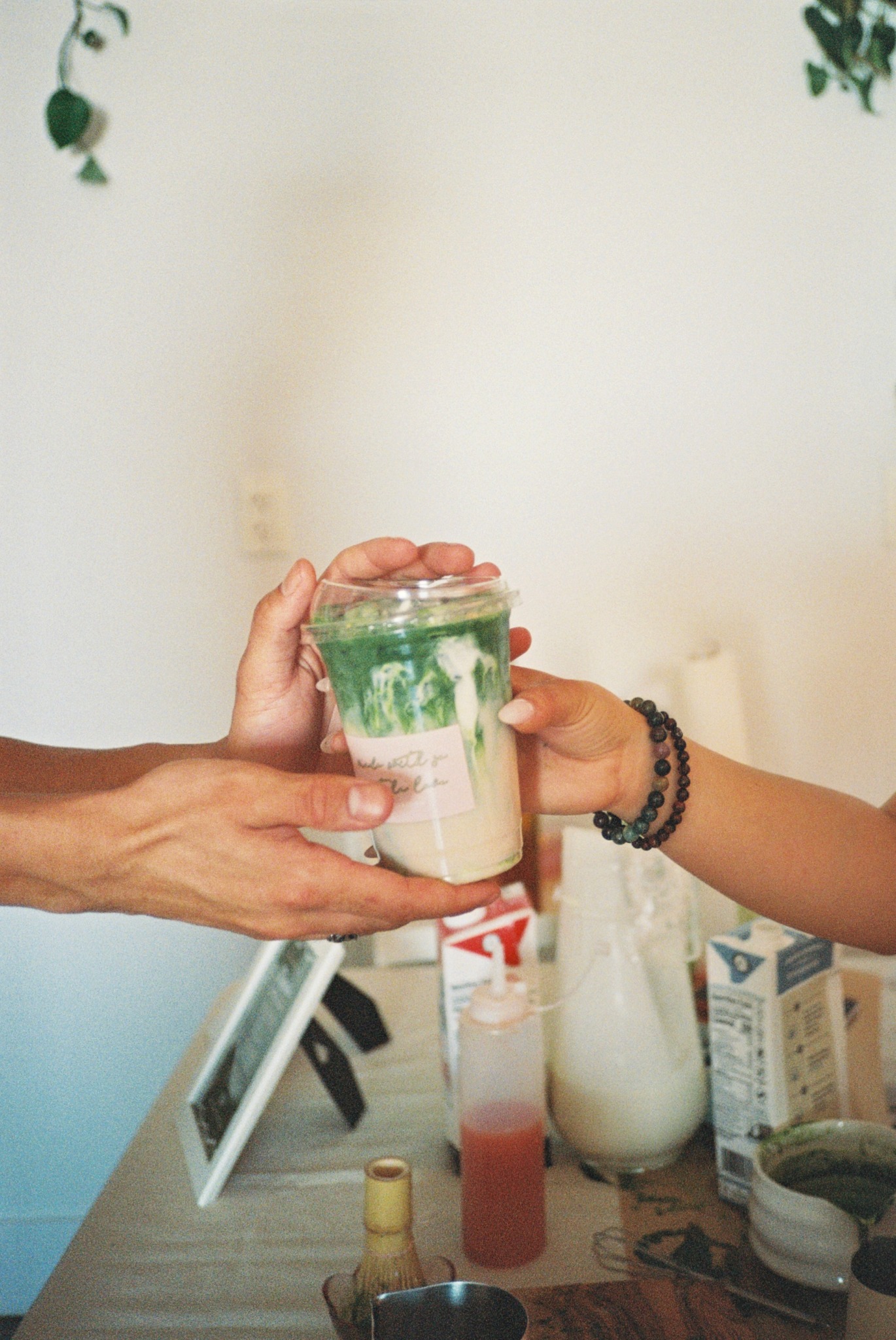 Two people exchange a layered drink with green and pink toppings in a cafe setting.