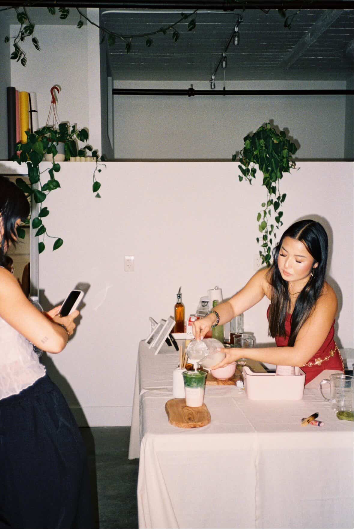Woman preparing food at a table with plants and kitchen items, another person taking a photo indoors.