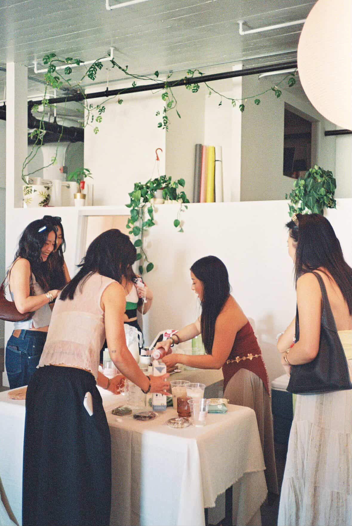 Group of women gathered around a table with drinks and snacks indoors, decorated with hanging plants and books.