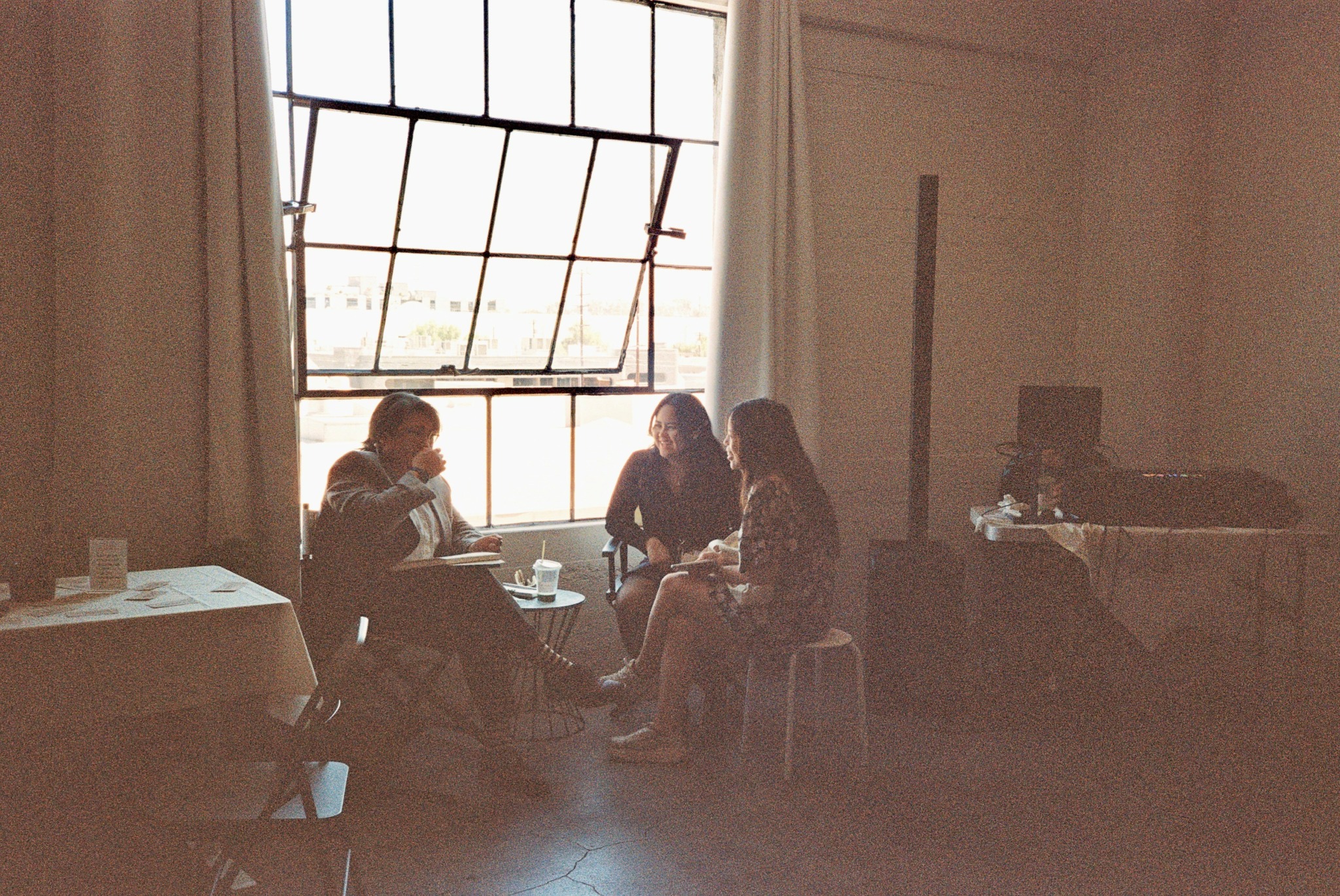 Four people sit around a table near a large window with open panes, in a room with white walls and curtains.