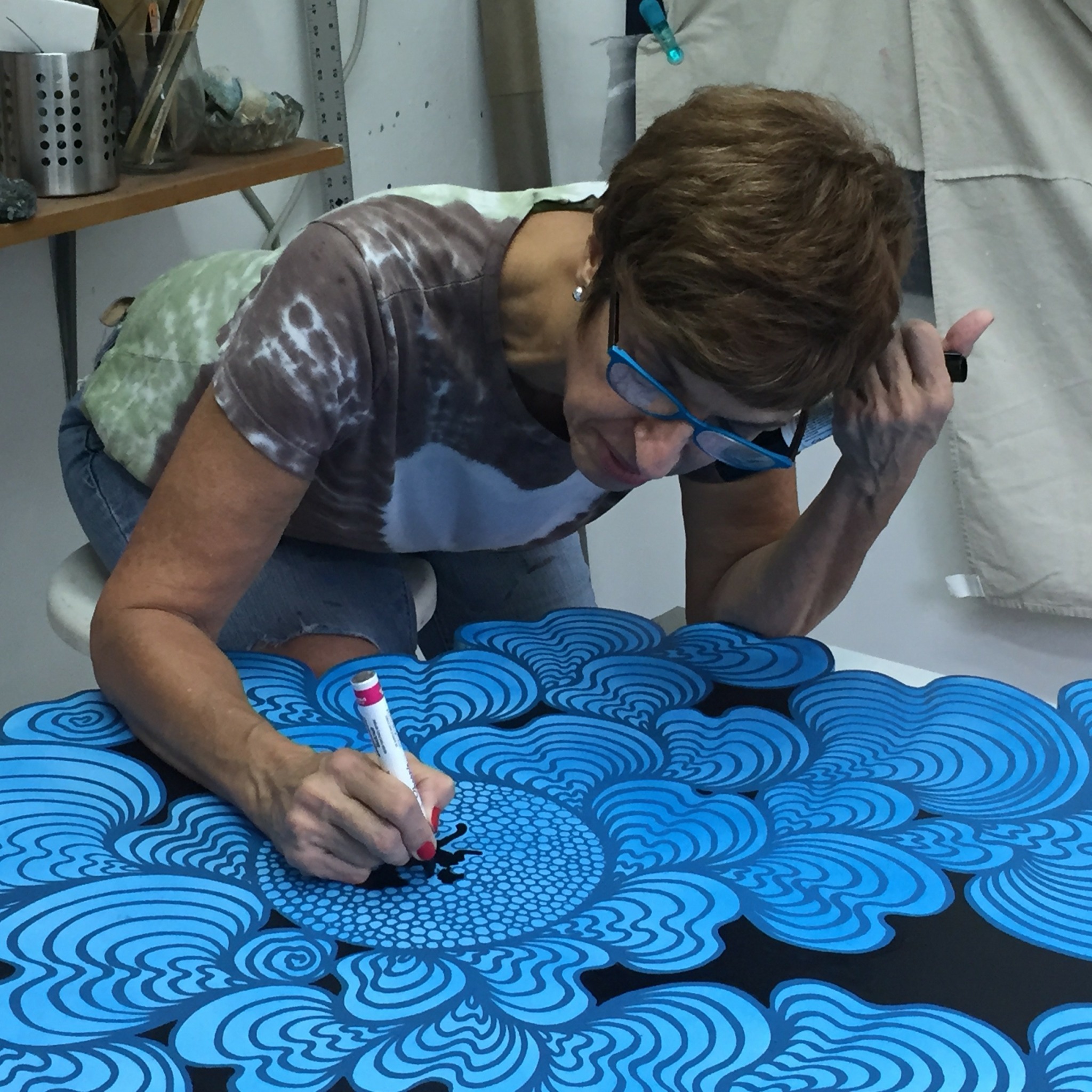 Woman with glasses drawing on a blue patterned surface in a room with shelves and curtains.