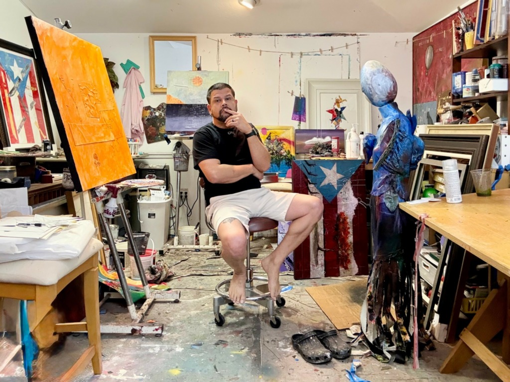 Man sitting on stool in art studio surrounded by paintings, sculptures, and art supplies.