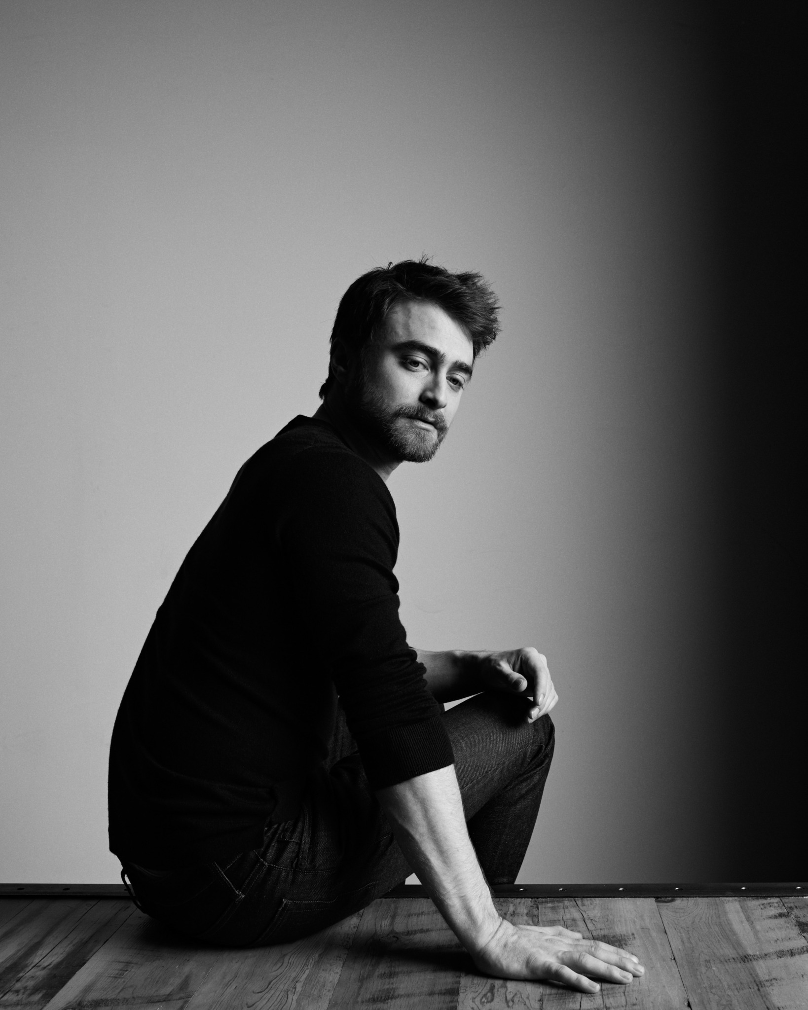 Man sitting on floor with one arm resting on his knee, looking at camera, in black and white.