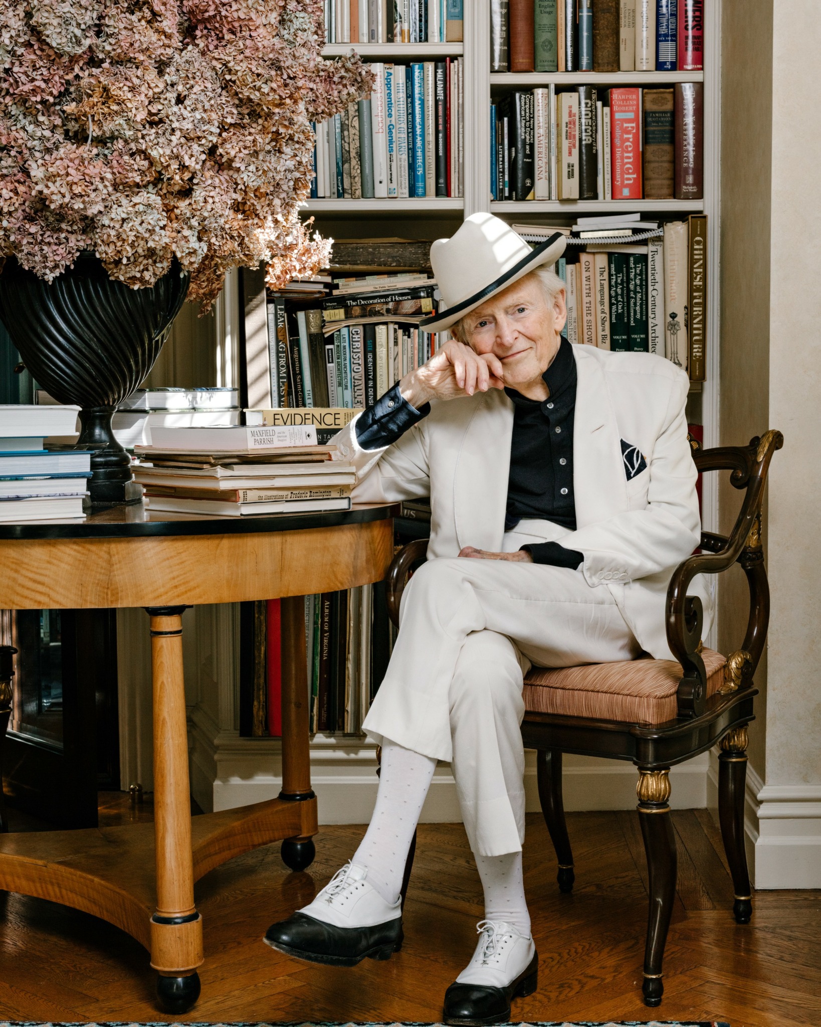 Elderly person sitting on a chair, wearing a white suit and hat, in a room with bookshelves and a large flower arrangement.