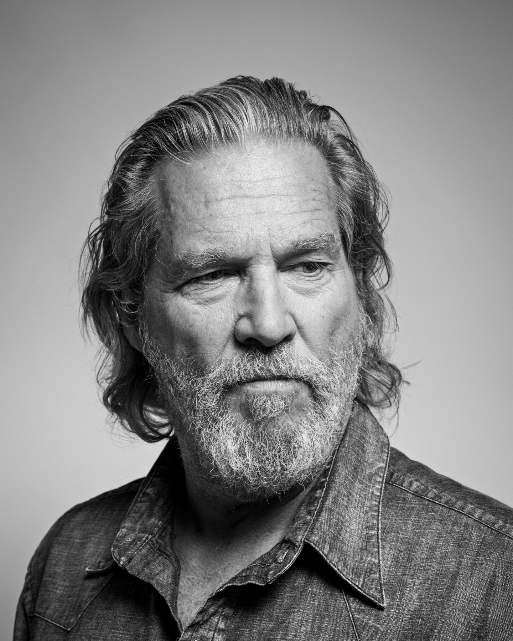 Black and white portrait of a middle-aged man with wavy hair and a beard, wearing a denim shirt.