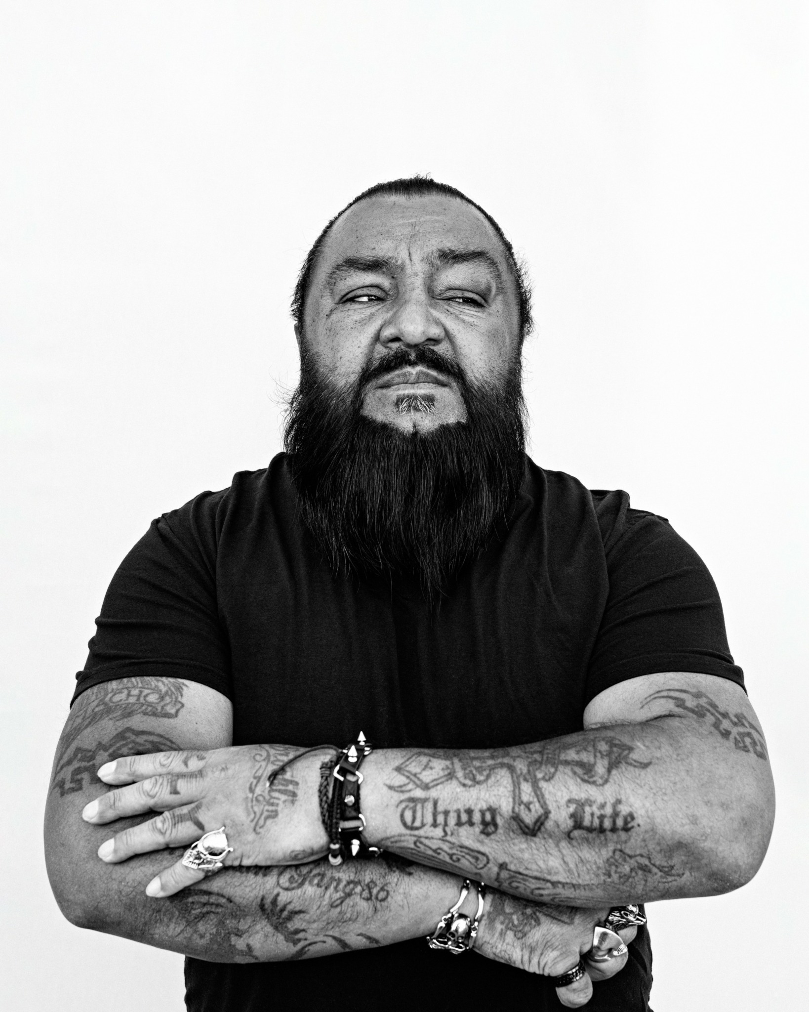 Man with a beard and tattoos crossing arms, wearing a black shirt, against a plain background.