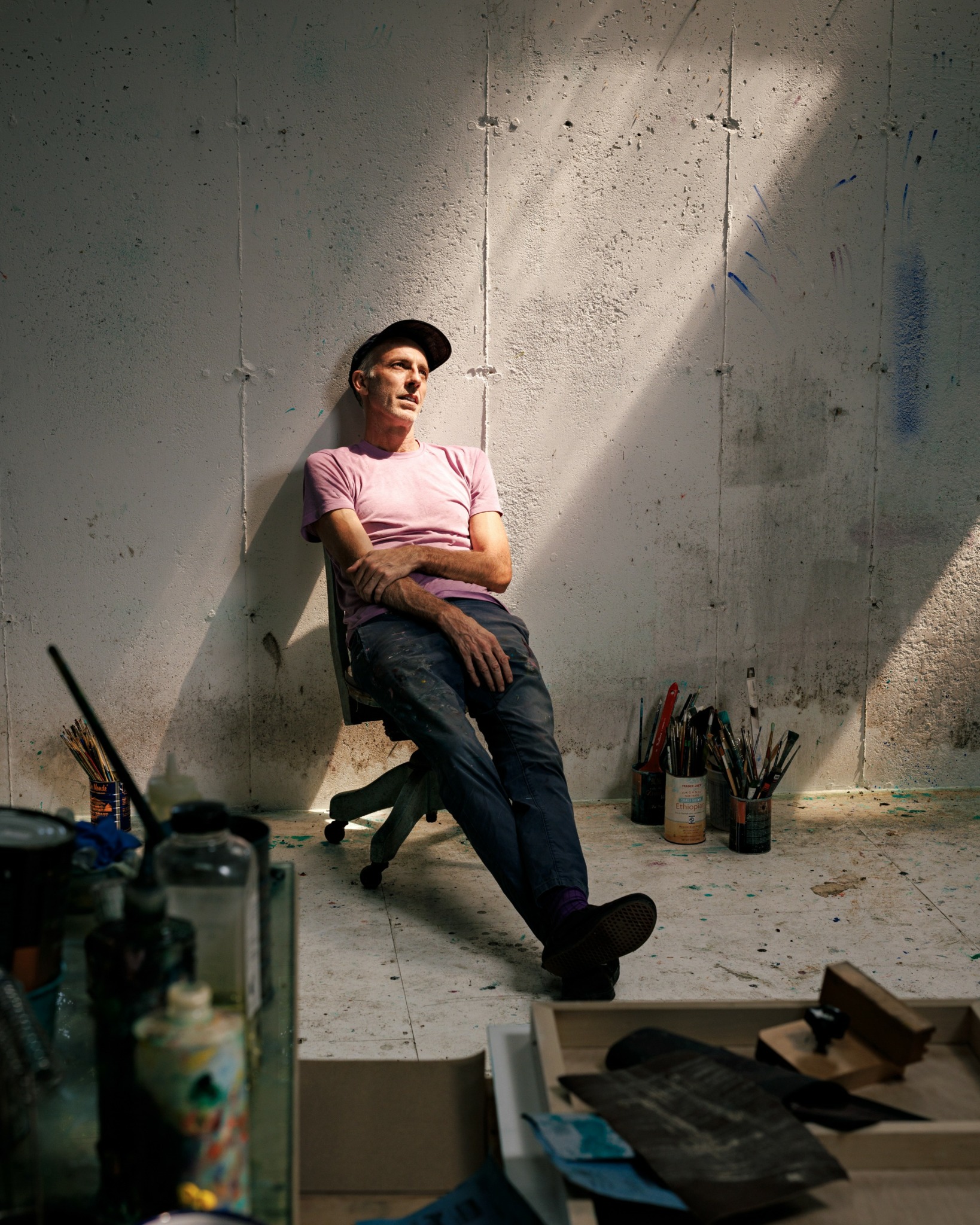 Person sitting on a chair against a concrete wall, illuminated by a diagonal beam of light, surrounded by art supplies.