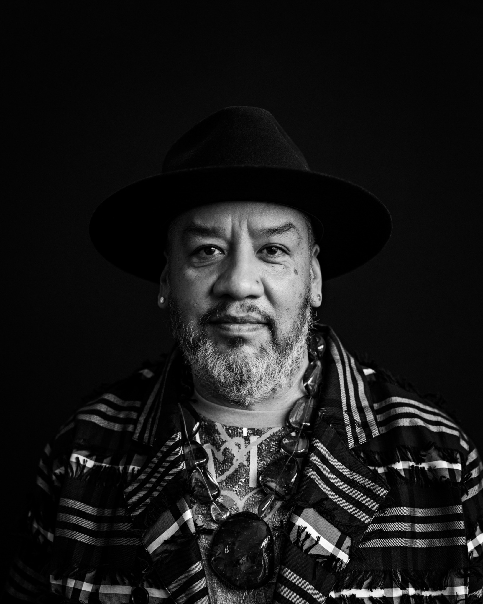 Man with a beard wearing a wide-brimmed hat and striped shirt, looking at the camera against a dark background.
