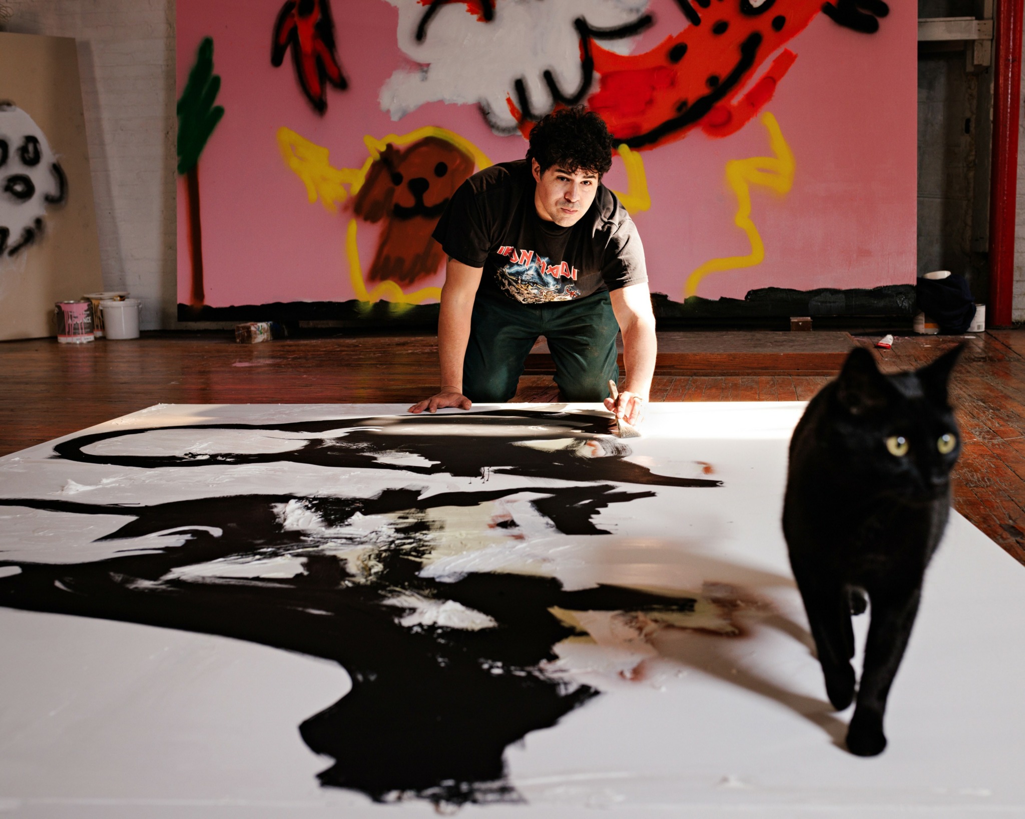 Person crouching on floor with large black and white painting, black cat walking nearby, colorful artwork on wall behind.