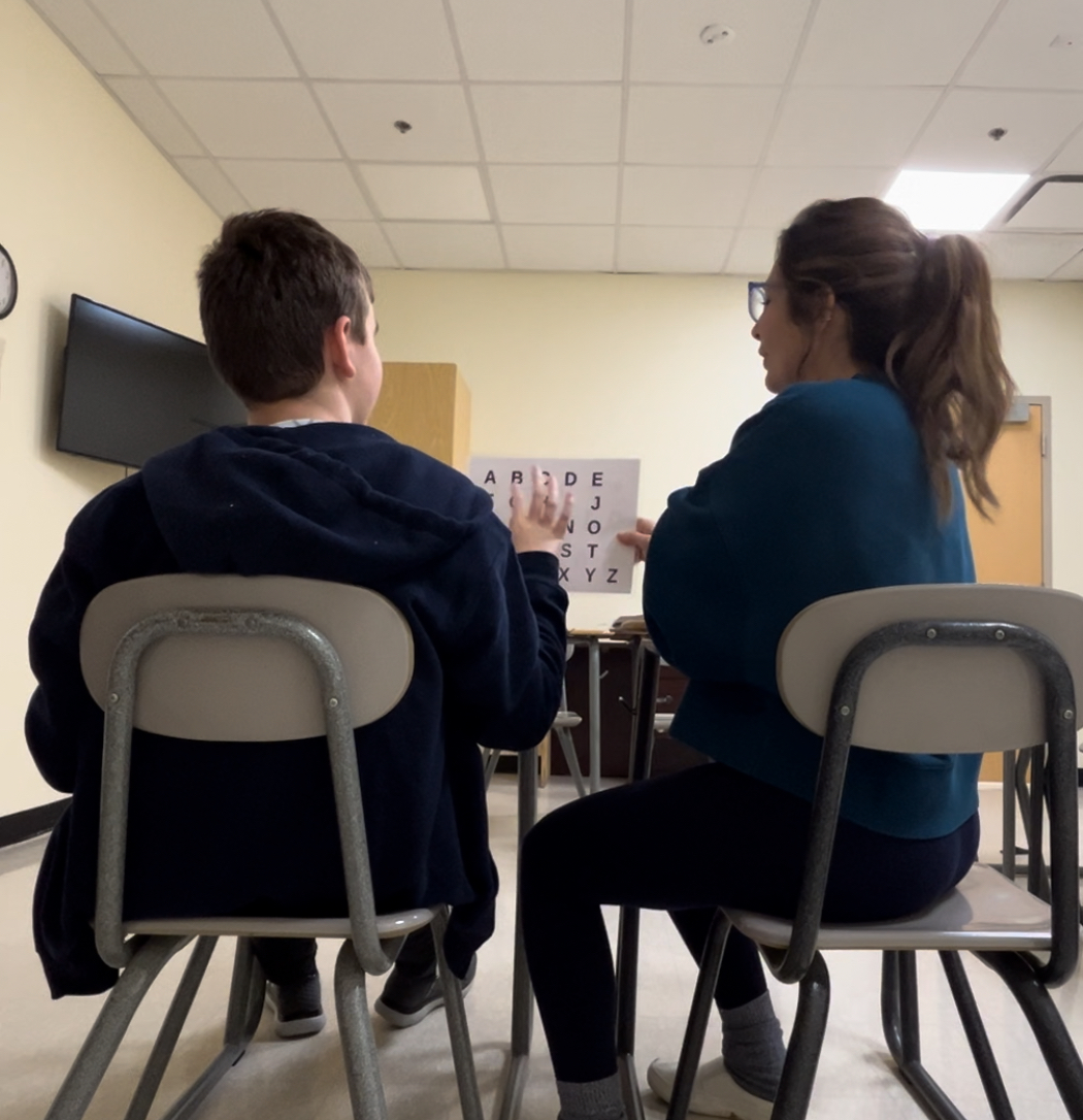 Two people sitting on chairs facing each other in a room with a television and an alphabet chart on the wall.