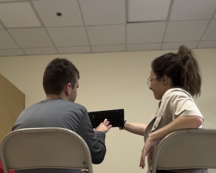 Two people sitting and exchanging a black object in a room with a ceiling grid.