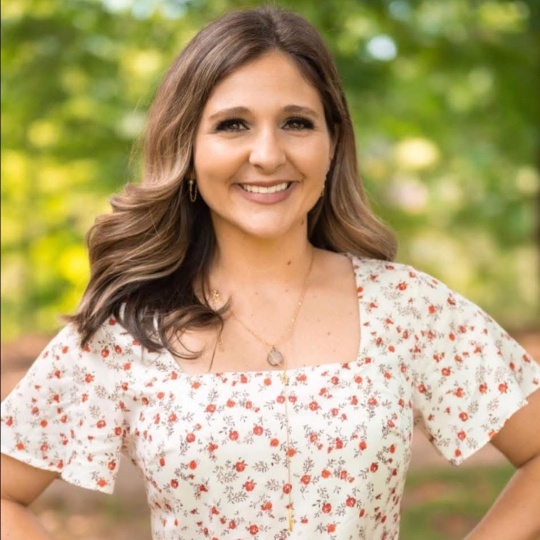 Smiling woman with wavy brown hair outdoors, wearing a floral dress and gold jewelry.
