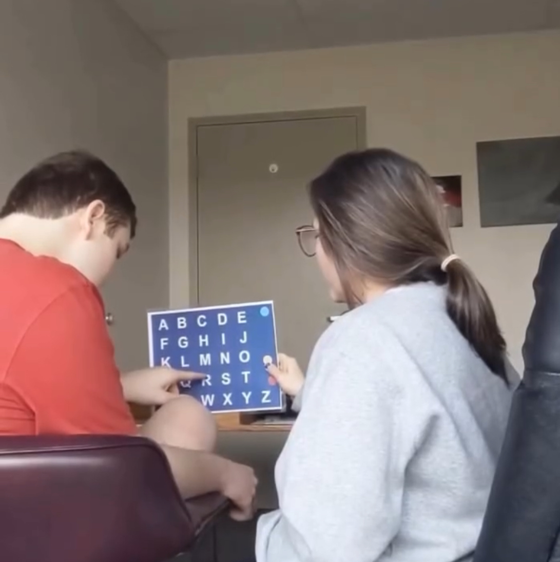 Two people sitting at a table, one pointing at a blue alphabet chart, the other wearing glasses and a gray sweater.