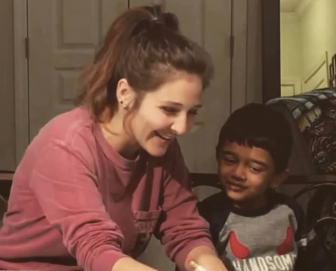 Young woman and child smiling and interacting indoors, woman wearing pink sweatshirt, child in gray shirt with red and white design.