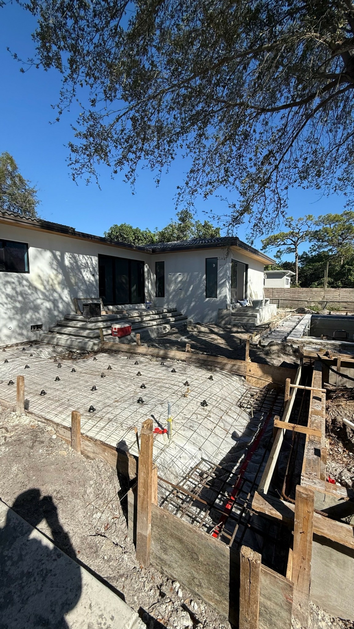 Construction site with rebar and wooden forms for concrete, house in background, trees and blue sky above.
