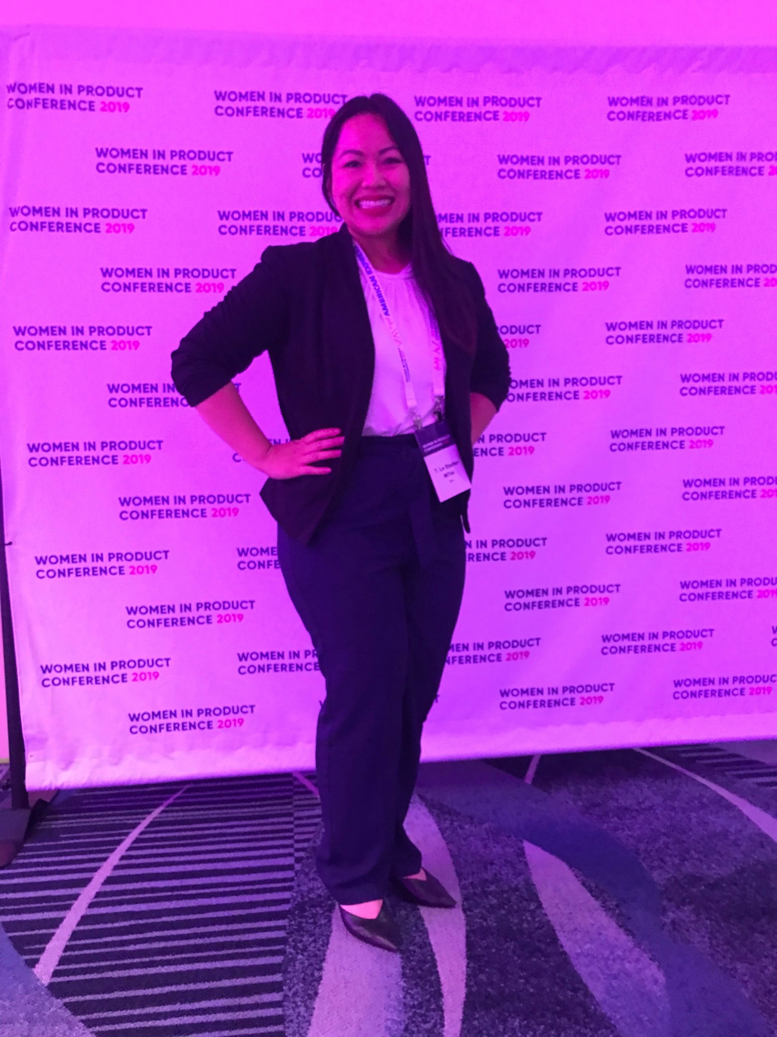 Woman standing on stage with a backdrop, smiling, wearing a blazer and pants, hands on hips, purple lighting.