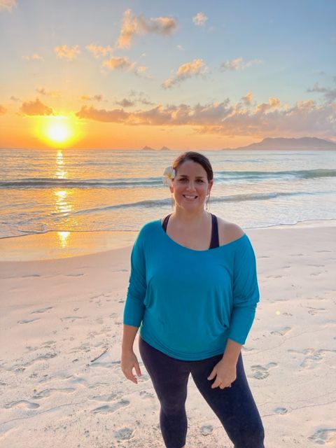Woman standing on beach at sunset, wearing blue top and dark pants, with ocean and sky in background.