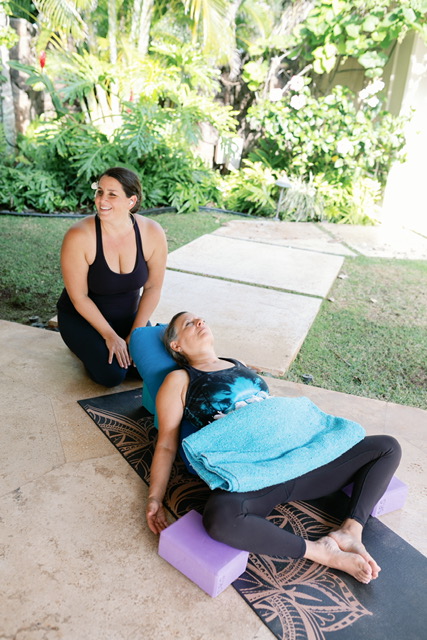Two women outdoors on yoga mats, one sitting and one lying down, surrounded by greenery.