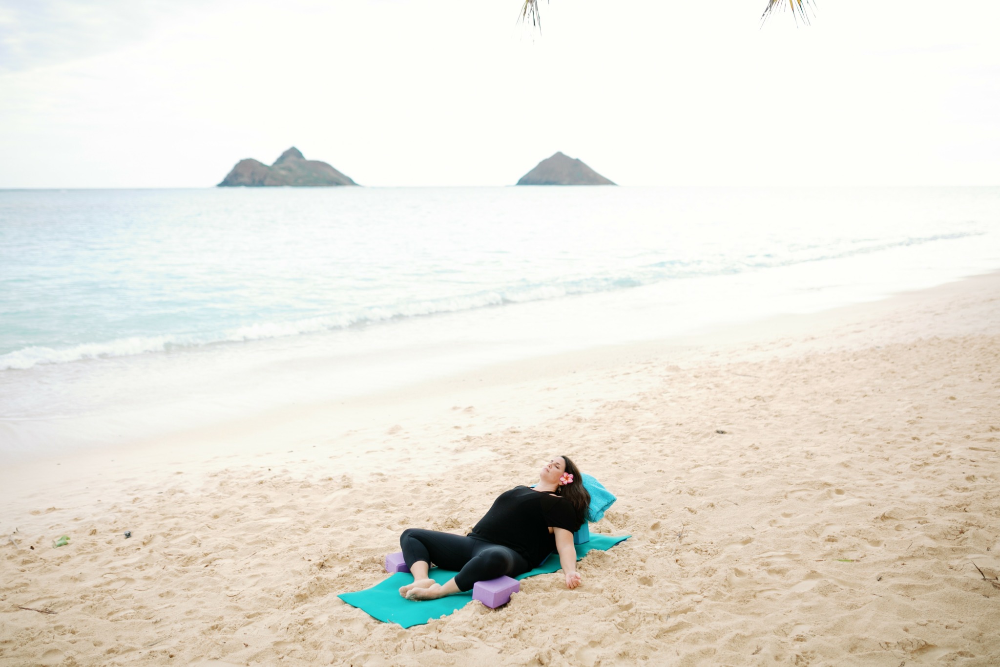Person lying on a beach towel with a purple ball, relaxing on sandy beach with ocean and islands in background.