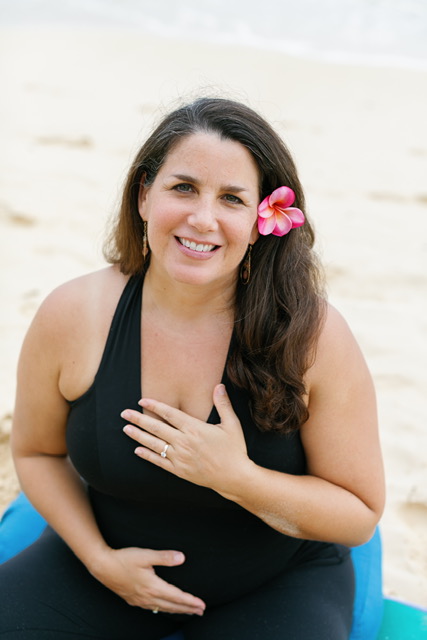 Woman with long dark hair and a pink flower in her hair, smiling on a beach, wearing a black top, hand on chest.
