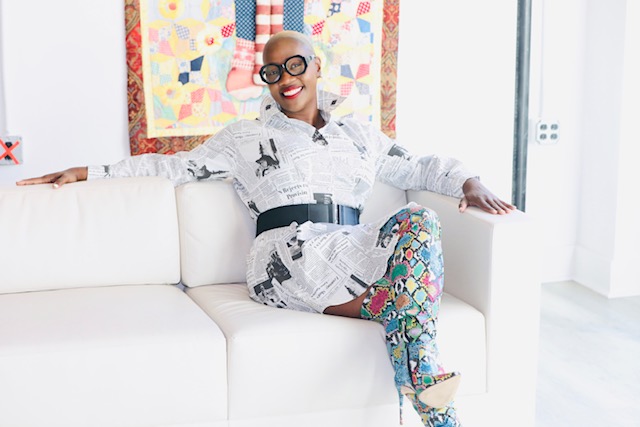 Person sitting on a white sofa, smiling, wearing glasses, a patterned dress, and colorful leggings.
