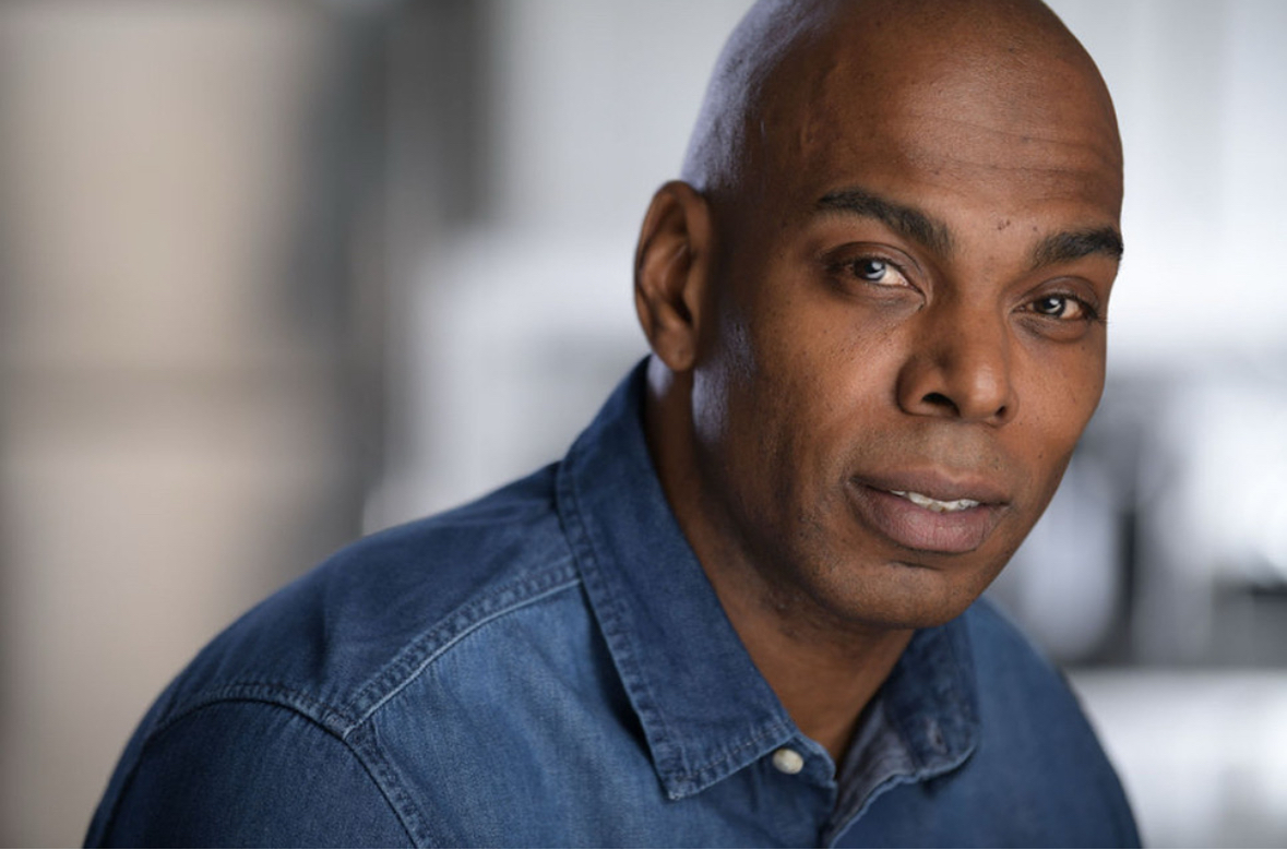 Portrait of a man with a shaved head wearing a denim shirt, looking at the camera.