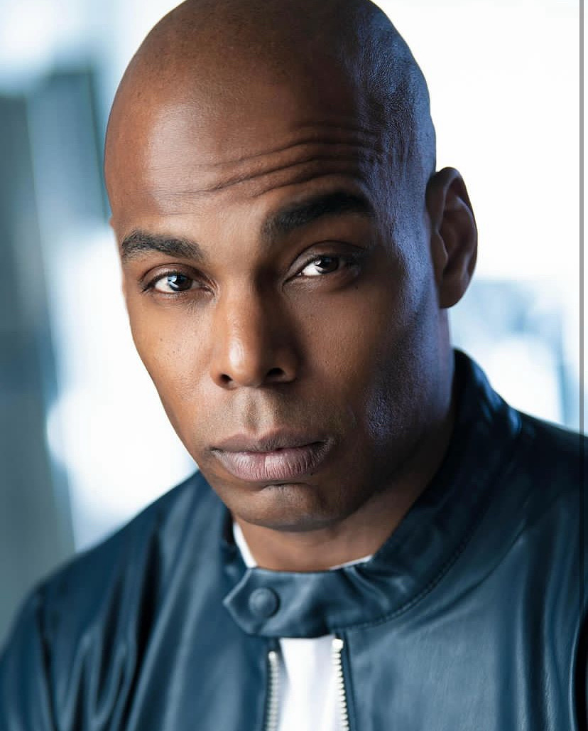 Close-up of a man with a shaved head wearing a dark jacket, looking directly at the camera.