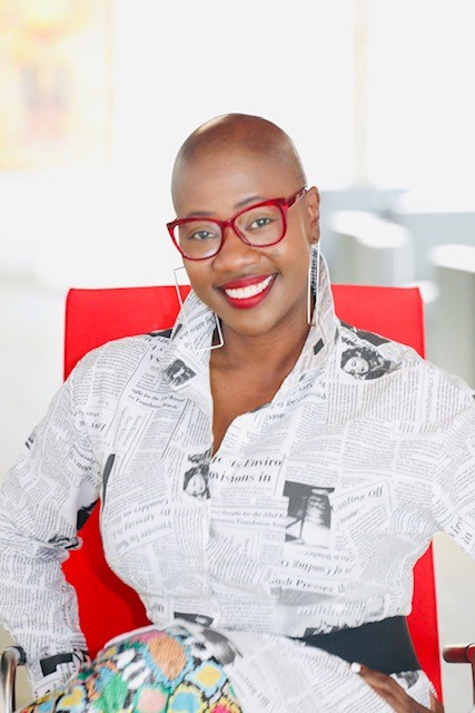 Woman with short hair, red glasses, and earrings, smiling, sitting on a red chair in a bright room.
