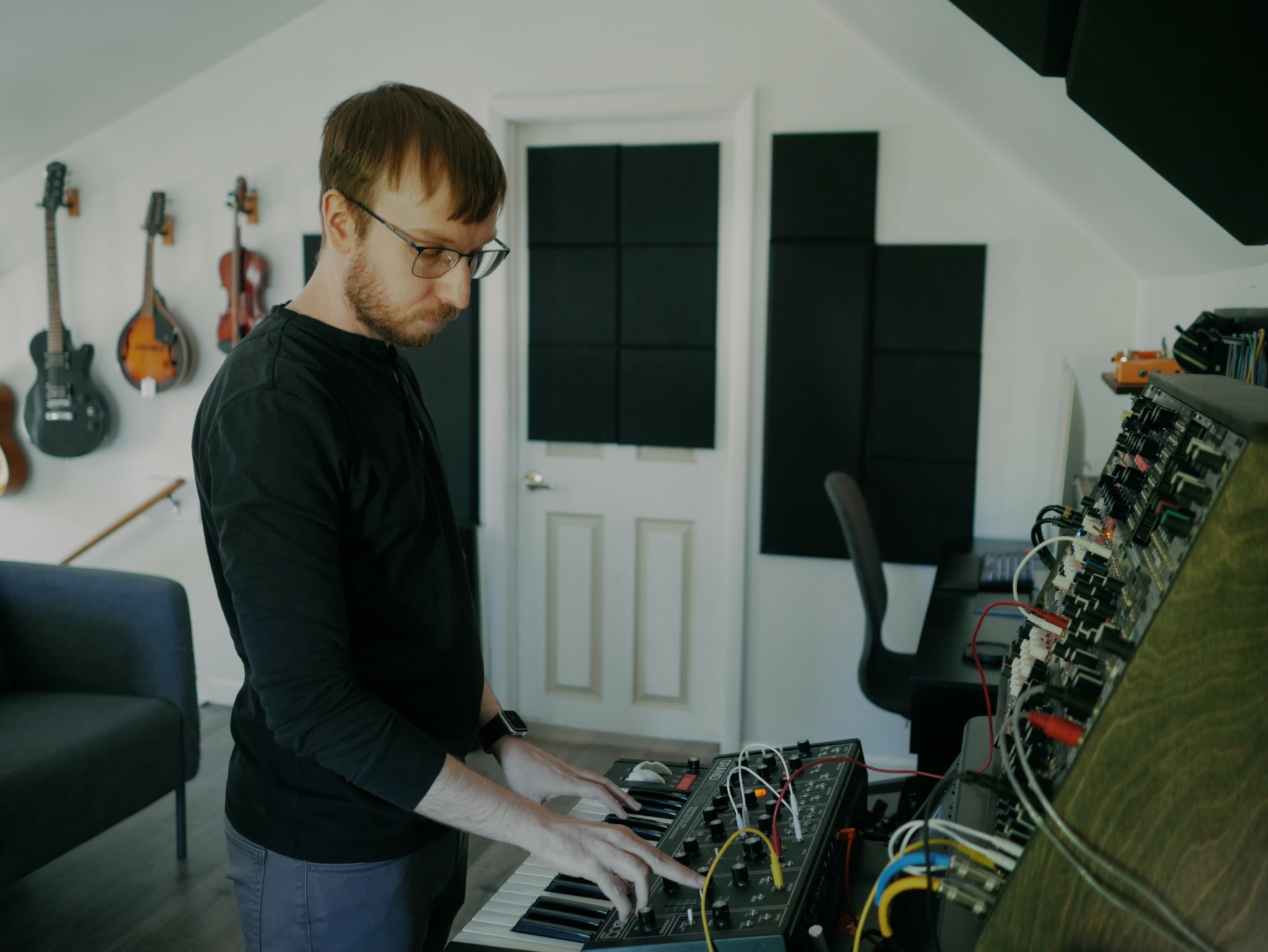 Man with glasses playing keyboard in music studio with guitars on wall and soundproof panels.
