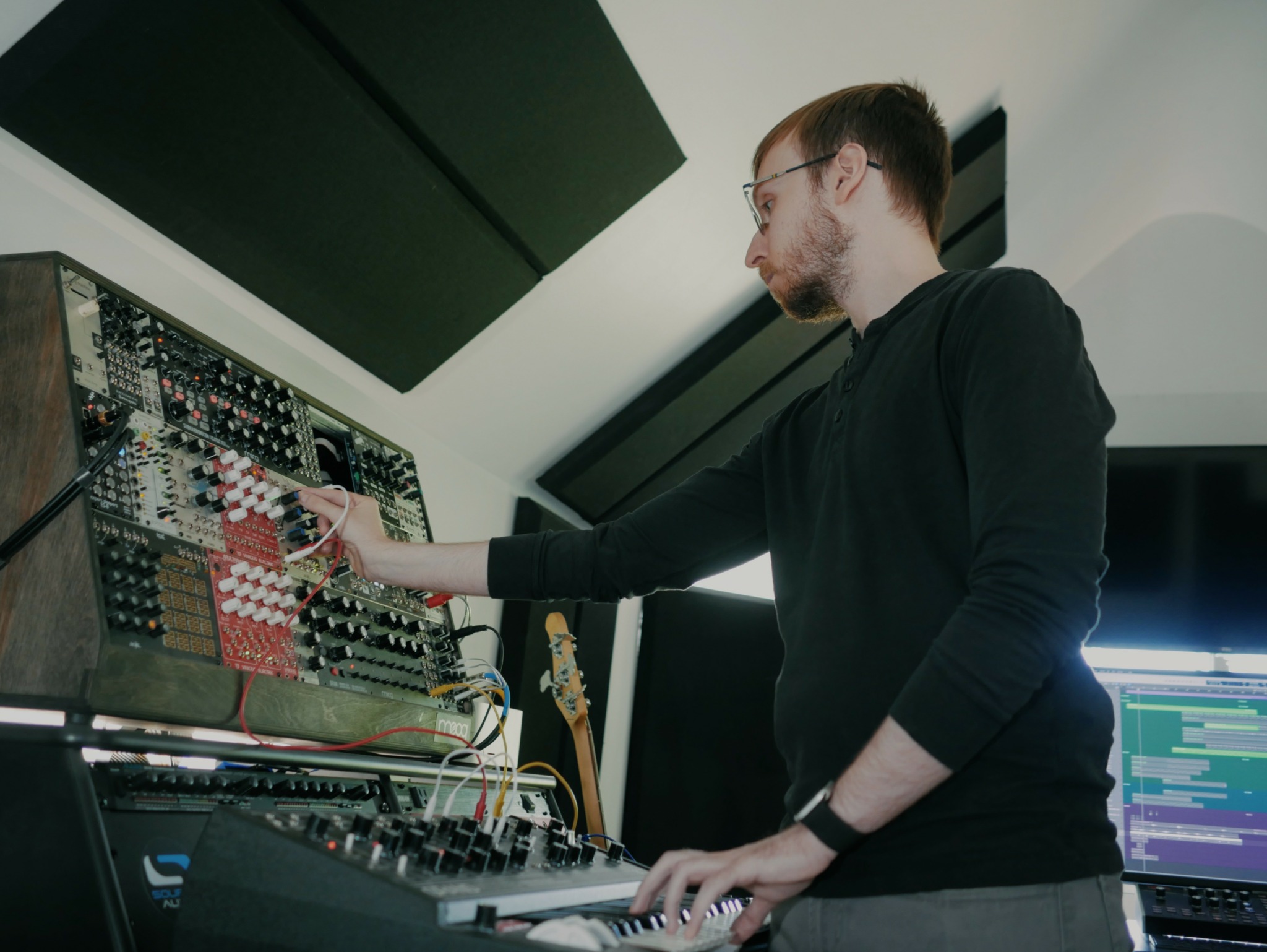 Man adjusting audio equipment in a room with computer screens and sound gear.