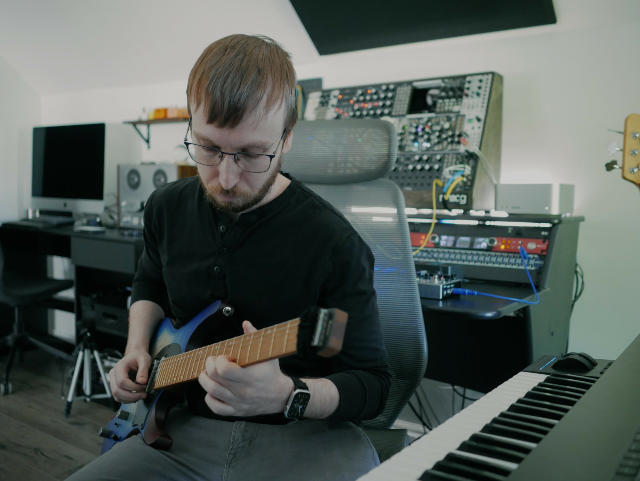 Person with glasses playing electric guitar in a music studio with equipment and keyboard.
