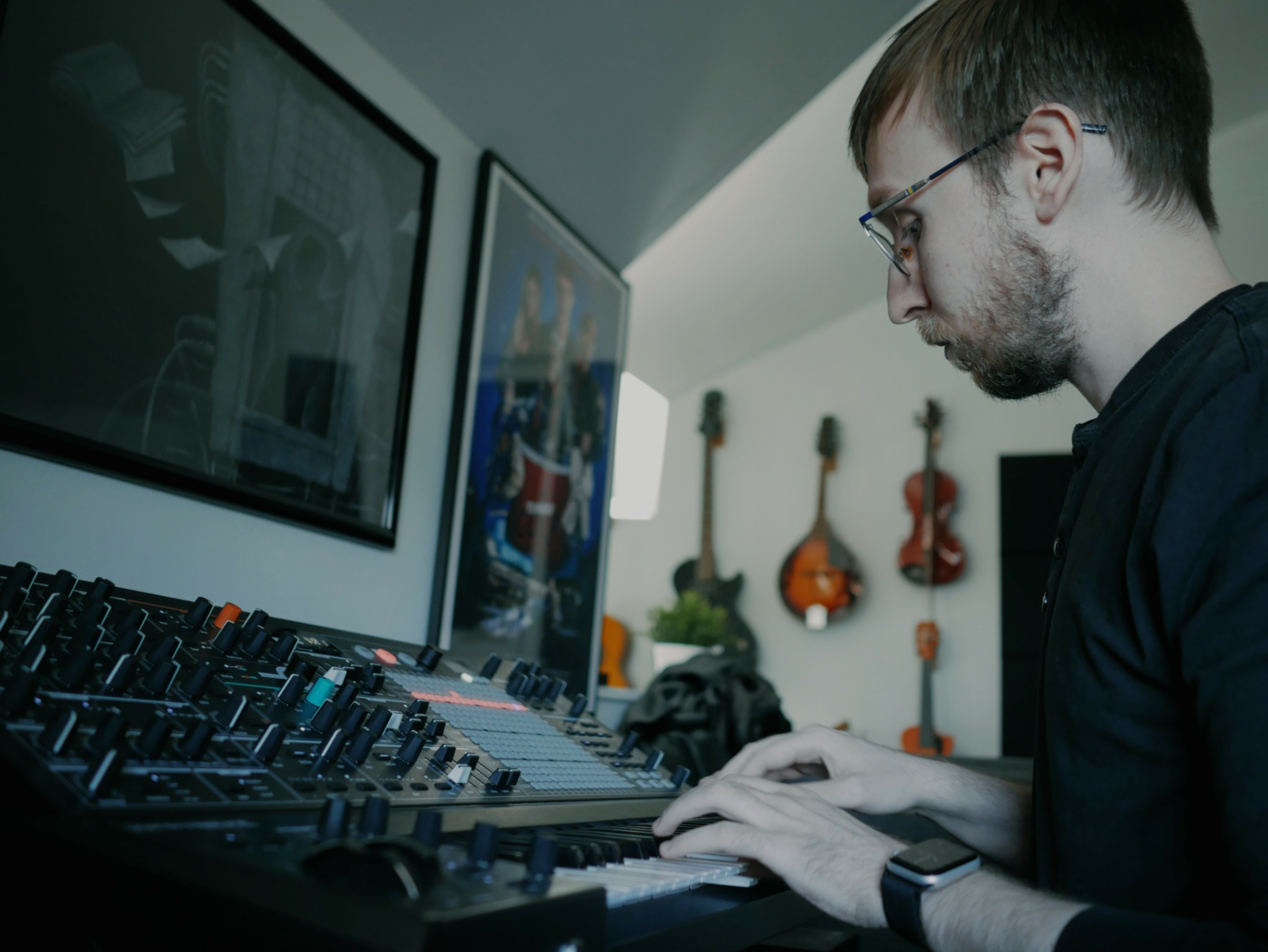 Person with glasses using a DJ mixer with monitors on wall and guitars in background.