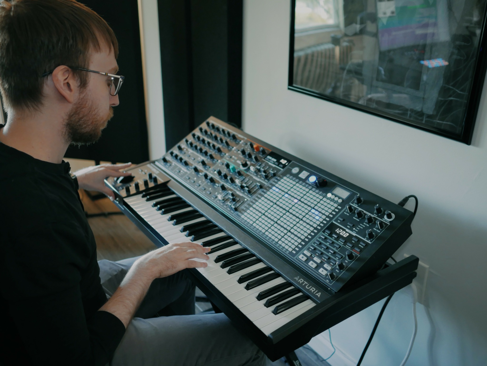 Person with glasses playing a synthesizer keyboard in a music studio, looking at a monitor screen.