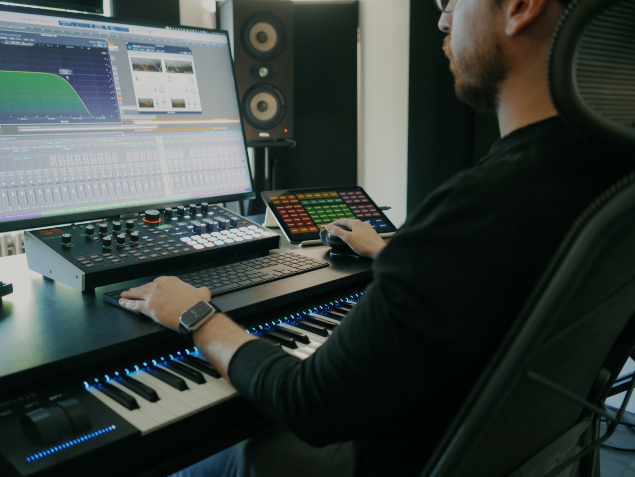 Person working at a music production workstation with a computer, keyboard, and colorful control pad.