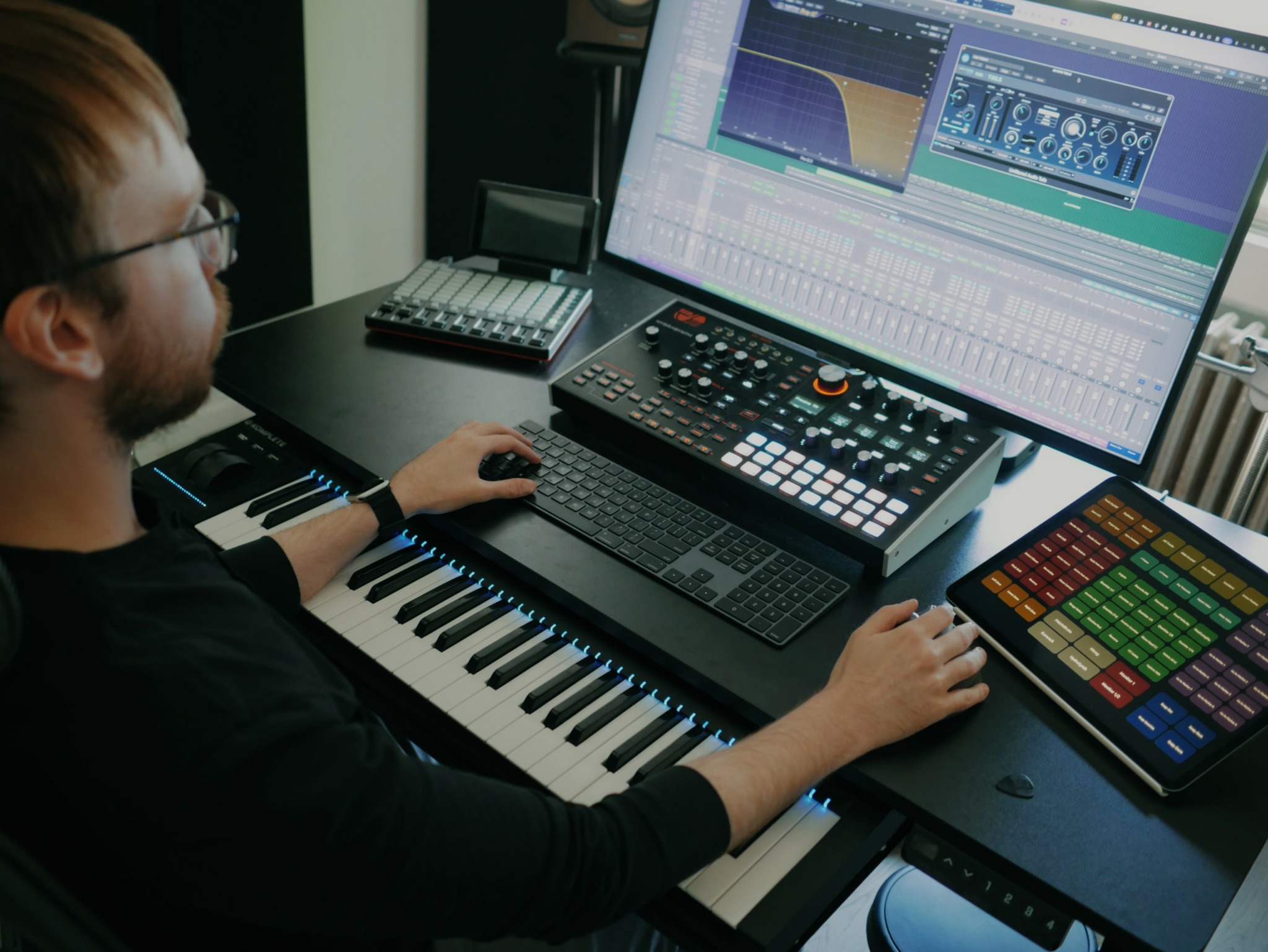 Person working at a music production setup with a computer, keyboard, and colorful pad controller.