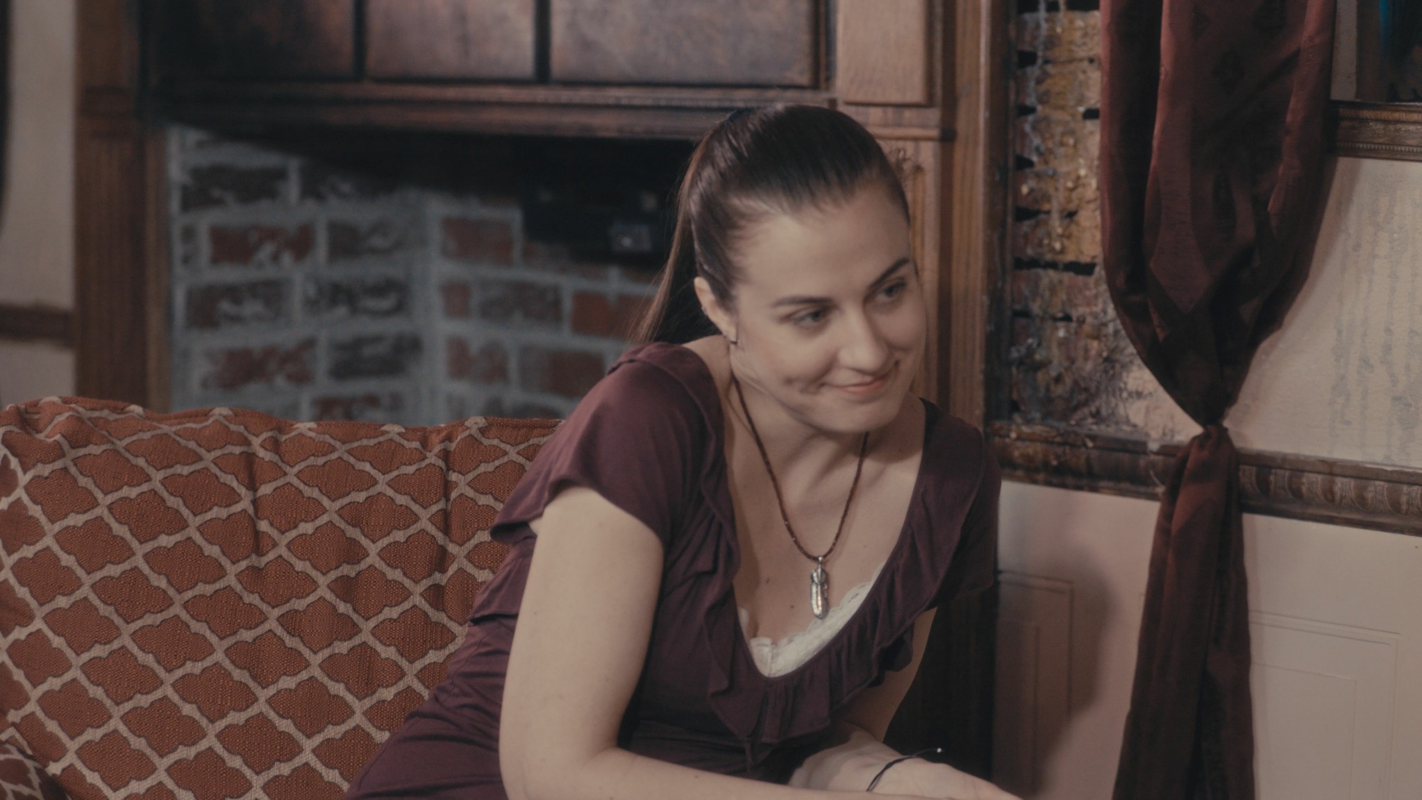Young woman with dark hair tied back, sitting on a patterned sofa in a room with brick and wood decor.
