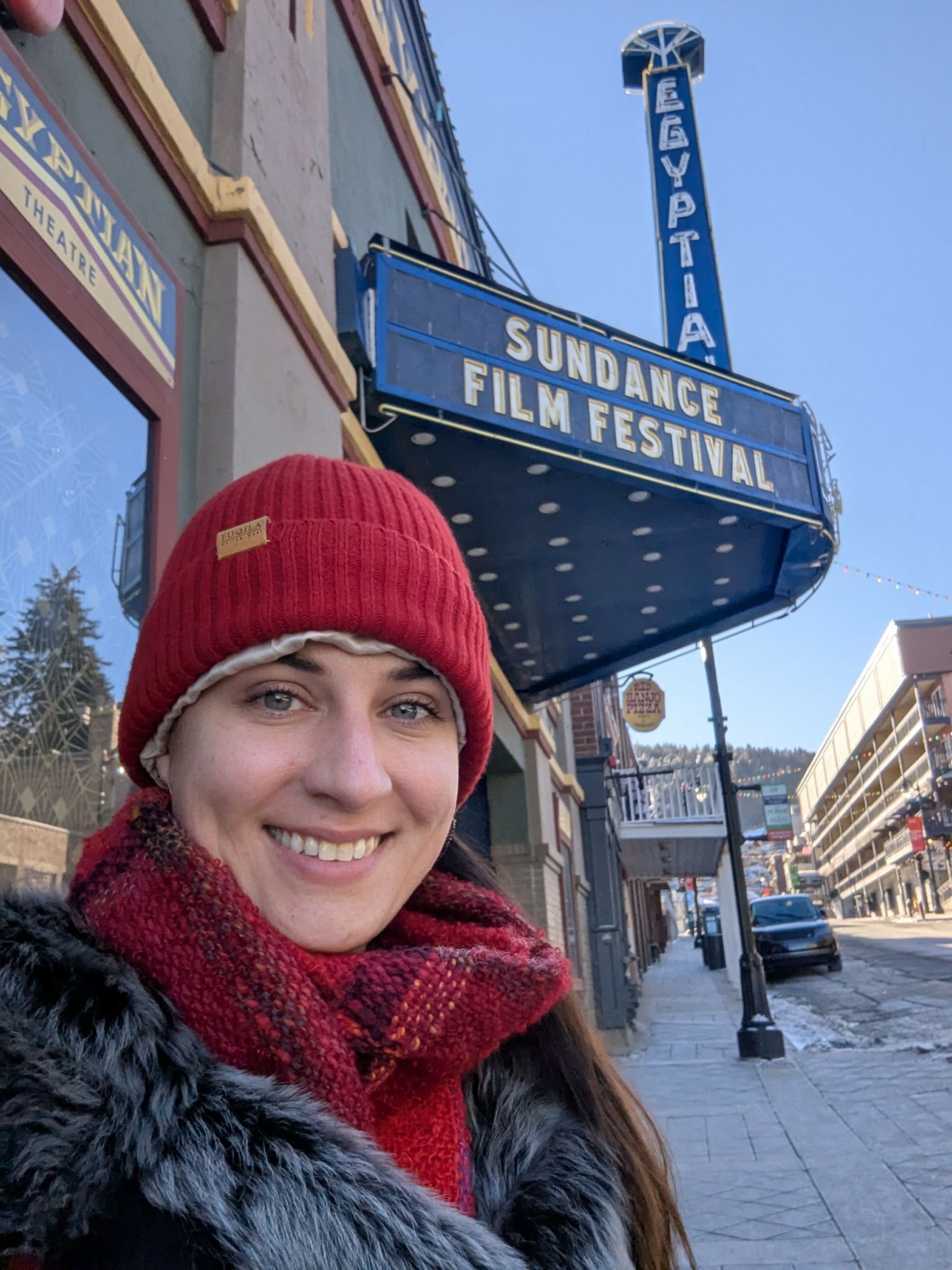 Smiling woman wearing red beanie and scarf standing on sidewalk near theater marquee.