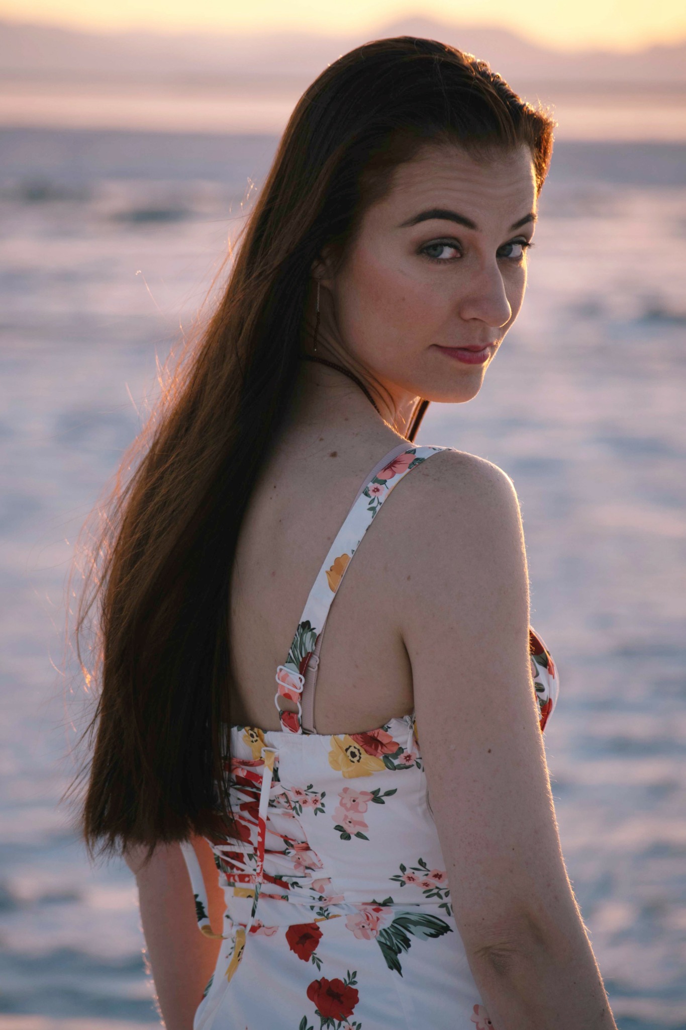 Young woman with long dark hair looking over shoulder at sunset beach, wearing floral dress.