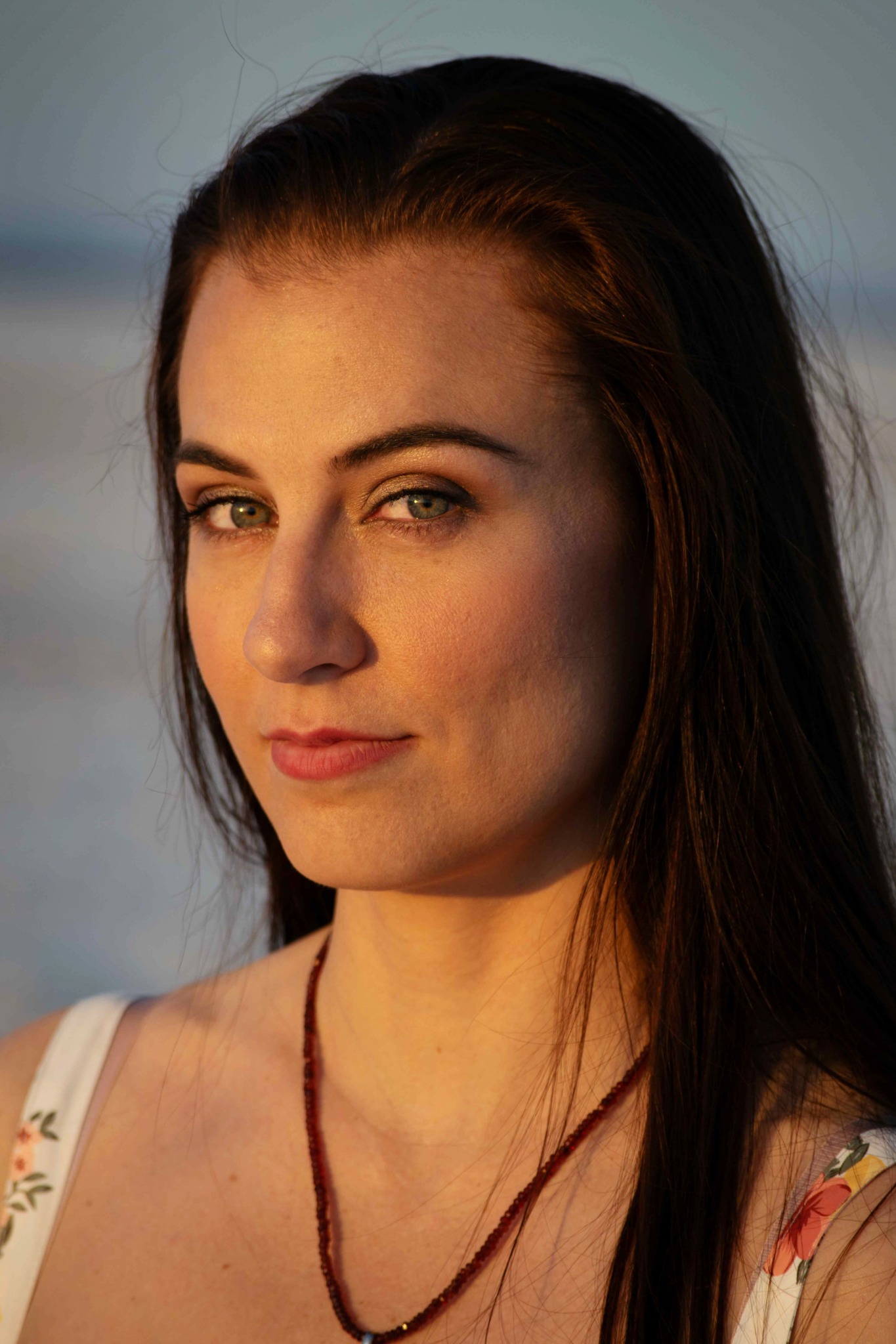 Close-up of a woman with long dark hair and blue eyes, looking at the camera outdoors during sunset.